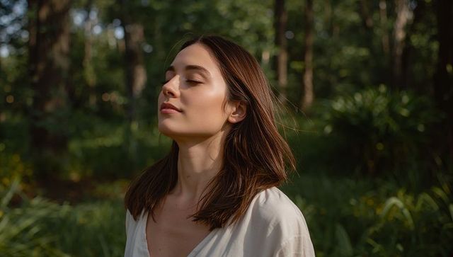 Young woman breathing and closing eyes in sunlit forest wearing v-neck blouse