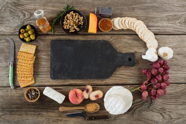 Assorted gourmet foods including cheese, crackers, grapes, nuts, honey, olives, garlic, and peaches arranged around an empty cutting board on a rustic wooden table. Ideal for use in culinary blogs, recipe websites, food magazines, and kitchen-related advertisements. Perfect for illustrating food preparation, gourmet dining, and rustic kitchen themes.