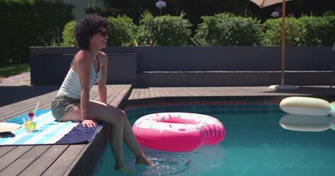 African american woman sitting poolside dipping feet in water with pink donut float