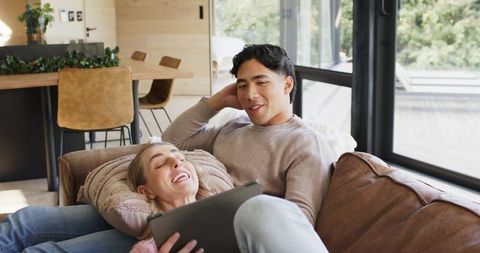 Multicultural couple lounging on leather sofa using tablet in modern cozy living room