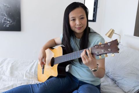 Mature woman practicing acoustic guitar on bed, relaxing with music at home