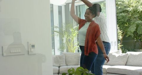 African couple dancing and twirling in sunlit living room by glass doors and modern sofa