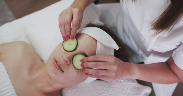 Caucasian woman lying back while beautician gives her a facial. putting cucumber slices on her eyes. customer enjoying treatment at a beauty salon.