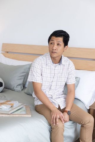 Contemplative mid-adult asian man sitting on bed edge in minimalist modern bedroom