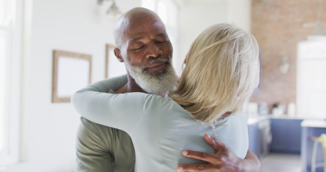 This image shows a senior couple from diverse backgrounds embracing warmly and showing affection in a cozy home setting. Ideal for use in projects promoting relationships, senior care, mental well-being, and emotional connection, as well as advertisements focusing on family life and comfort.