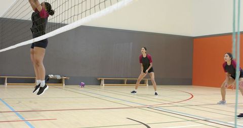 Diverse women volleyball team jumping and defending at net during indoor training session
