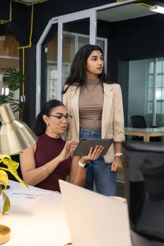 Two professional women collaborating on tablet and laptop in modern office workspace