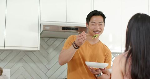 Asian man offering bowl to partner while smiling and sharing food in modern kitchen