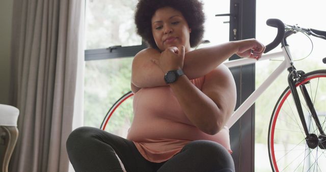 This image depicts a woman stretching her arm muscles while sitting cross-legged on the floor next to a bicycle in her home. She appears focused and determined, making it a perfect representation of fitness and healthy lifestyle. Ideal for use in articles, blogs, or ads promoting home workouts, fitness routines, well-being practices, or athletic wear.