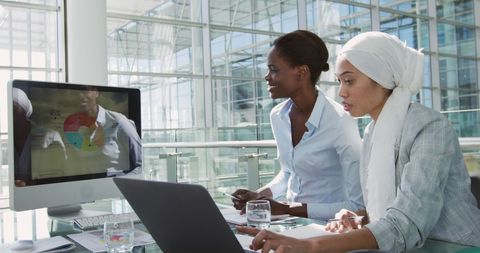 Two businesswomen, one of whom is African American and the other biracial, are finding solutions together in their vibrant office. They are working together at computers with graphs and data, elucidating the concepts of teamwork and workplace diversity. This visual can work well for promoting multicultural office environments, teamwork development, human resources materials, or diversity and inclusion campaigns.