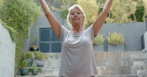 Senior woman practicing morning stretch on garden steps with raised arms in courtyard