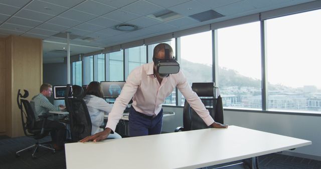 A man wearing a VR headset stands in a modern office, introducing a technology-driven work environment. In the background, other employees are working at their desks on computers. This image is perfect for businesses, tech blogs, and articles related to innovative office environments, virtual reality applications in business, and technological advancements in the workplace.