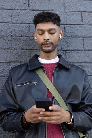 Young indian man using smartphone with earbuds wearing leather jacket and crossbody strap