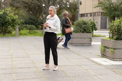 Mature diverse women standing in city plaza checking phone holding coffee with suitcase
