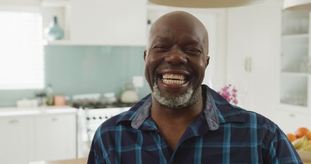 Mature African American man in a modern, bright kitchen. He is wearing a plaid shirt and is smiling warmly, creating a sense of happiness and contentment. Perfect for use in lifestyle, home, and positive emotion concepts, as well as advertisements promoting happiness and comfortable living environments.