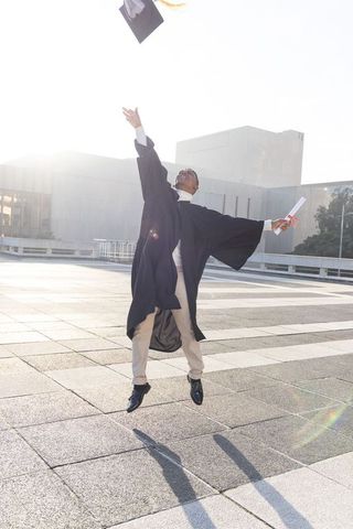 African american graduate jumping and tossing cap while holding diploma on urban campus