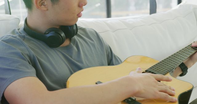 Asian male teenager with headphones playing guitar and sitting in living room. spending time alone at home.