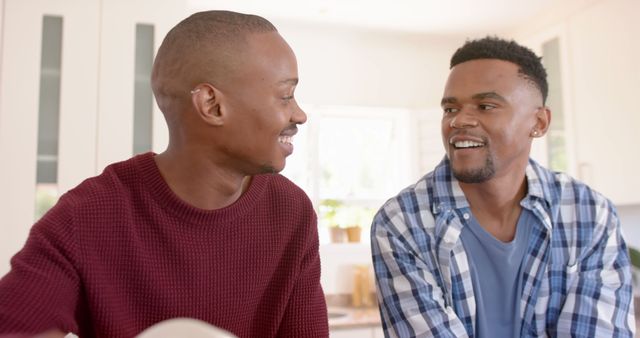 This image depicts two young men smiling and having a cheerful conversation in a bright, sunny kitchen, illustrating the themes of friendship, joy, and casual interaction. Perfect for promoting social connections, lifestyle blogs, mental health awareness, and advertisements aimed at fostering a positive, relaxed atmosphere.