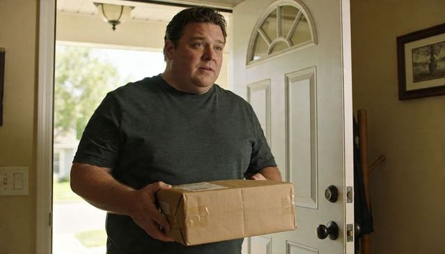 Middle-aged man holding cardboard package standing at open front door of suburban home