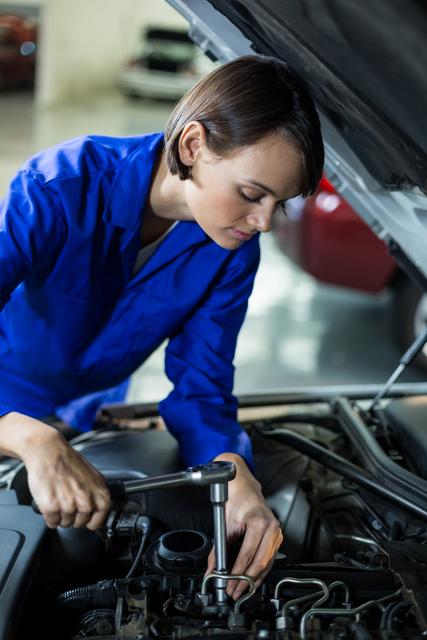 A female mechanic in a blue uniform is working on a car engine in a repair garage. This image is suitable for illustrating concepts related to automotive service, gender diversity in technical fields, and professional vehicle maintenance. It can be used for websites, articles, or advertisements focused on car repair services, mechanic training programs, or promoting women in STEM careers.