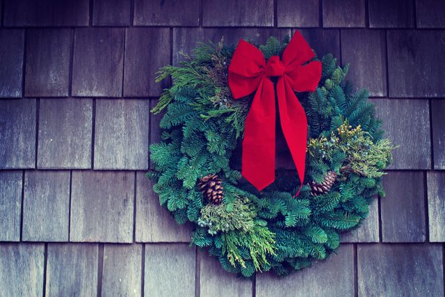 A festive holiday wreath adorned with pine cones and a large red bow hangs on a rustic wooden wall. This classic Christmas image is perfect for seasonal promotions, holiday greeting cards, advertisements, or decorating ideas.