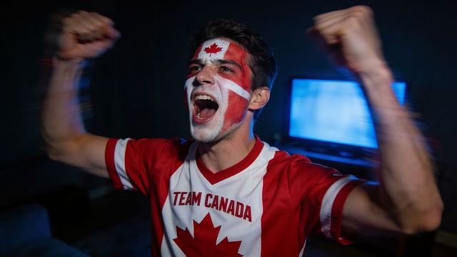 Cheering team canada fan wearing face paint and jersey raising fists in living room near tv