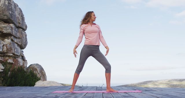 Woman is performing yoga on a wooden platform positioned in a rocky outdoor environment. She is standing on a pink yoga mat while stretching her arms and legs, promoting a sense of tranquility and fitness. Useful for content related to outdoor exercise, yoga practice, fitness and wellness, nature activities, or healthy living.