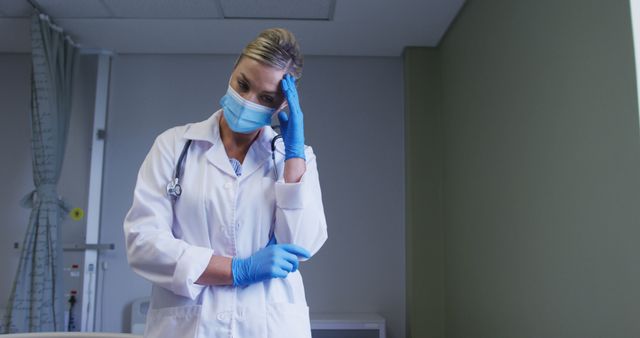 Thoughtful caucasian female doctor face mask and surgical gloves standing at hospital. medical healthcare during coronavirus covid 19 pandemic concept