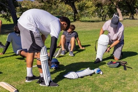 Cricket teammates adjusting leg pads on sunlit grass preparing for friendly outdoor match