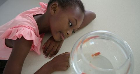 Young girl leaning on counter gazing at goldfish in glass bowl, serene childhood moment