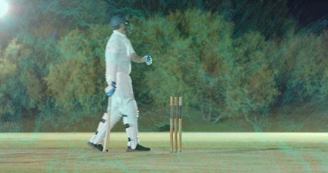 Cricketer walking past stumps on night pitch carrying bat wearing white pads