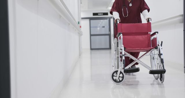 Healthcare worker wearing red scrubs pushing empty red wheelchair down hospital hallway. Ideal for medical and caregiving concepts, healthcare infrastructure illustrations, and imagery representing patient care and hospital environments.