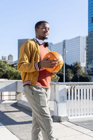 African american man holding basketball on urban rooftop with headphones and mustard jacket