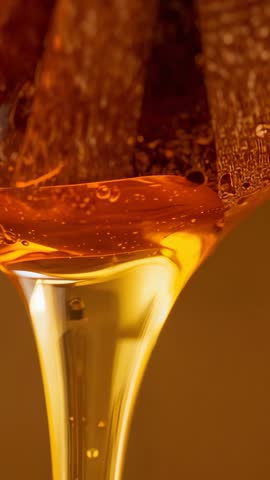 Vertical close-up of honey pouring from wooden dipper and flowing into glass slow motion
