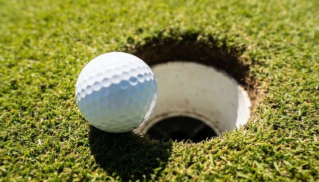 Golf ball teetering on cup edge on sunlit putting green close-up with shadow
