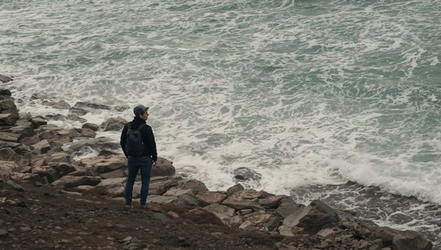 Man standing on rugged rocky coastline overlooking churning ocean waves and foam