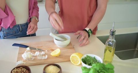 Friends preparing fresh herb pesto on bright kitchen countertop with lemon and olive oil