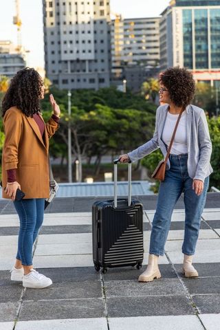 Female friends greeting with luggage on urban plaza tiles, casual blazers and jeans