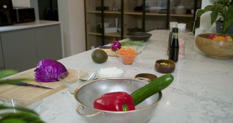 Holding red pepper and cucumber in metal colander on marble kitchen island with prep bowls