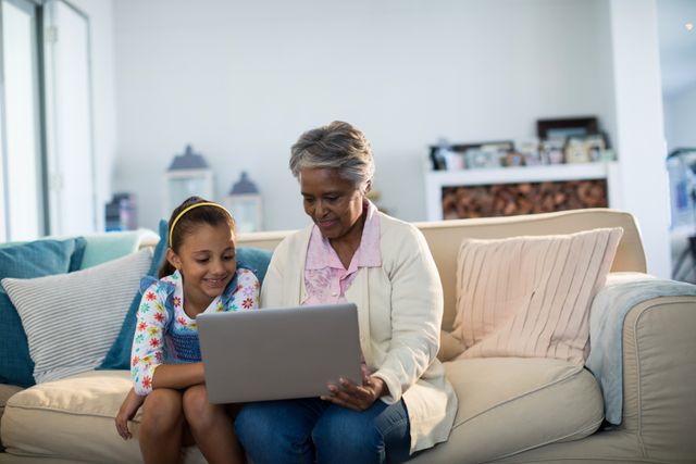 Grandmother and granddaughter sitting on a couch, using a laptop together. Perfect for illustrating family bonding, technology use across generations, or educational activities at home. Ideal for websites, blogs, or advertisements focusing on family life, senior and child interactions, or home learning environments.