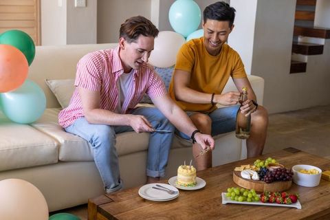 Two male friends celebrating at home cutting mini cake and popping sparkling bottle