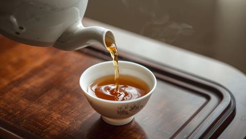 Porcelain teacup receiving golden-brown tea pouring from teapot on wooden tray