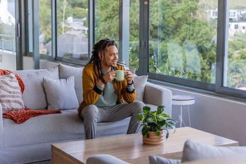 African american man with dreadlocks relaxing on sofa holding coffee by sunlit window