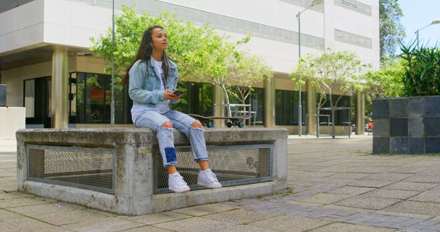 Young woman sitting outside on a campus in front of a modern building. She holds a smartphone and is dressed in casual denim attire, featuring a denim jacket and ripped jeans. Various trees and natural sunlight contribute to a serene, collegiate atmosphere. May be used for themes related to education, college life, relaxation, and casual fashion.