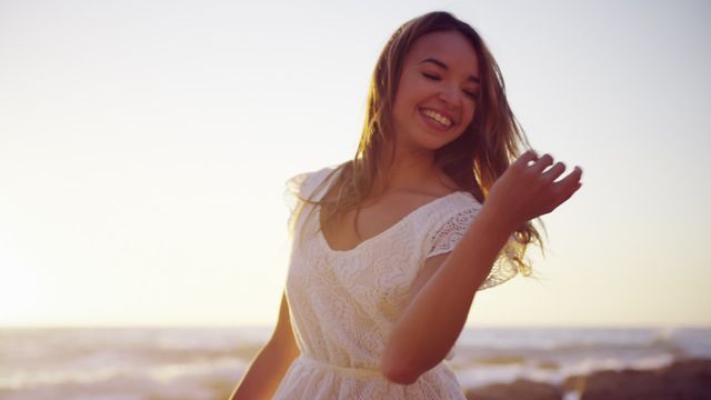 A young Caucasian woman is joyfully dancing on a beach at sunset, wearing a white dress. The warm light of the sunset and the ocean waves create a serene backdrop, highlighting a sense of freedom and happiness. Ideal for use in lifestyle and travel promotions, advertisements, or editorial pieces that focus on joy and enjoying life by the sea.