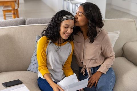 Diverse mother kissing teenage daughter while reading happy letter on sofa