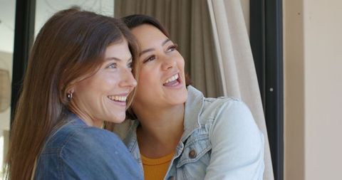 Smiling diverse friends embracing by window, denim jacket and mustard top, natural light