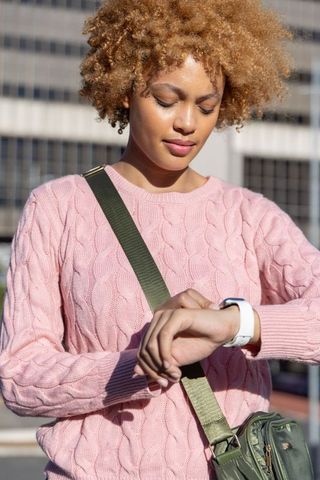 African american woman checking smartwatch outdoors in pink cable knit sweater urban plaza