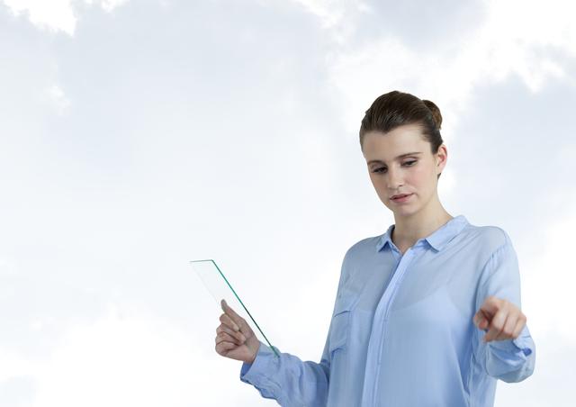 A professional woman in a light blue blouse interacts with an augmented reality interface. She holds a transparent glass tablet in one hand while using her other hand to touch or manipulate the digital space around her. The bright clouded background adds a futuristic feel. This image can be used in technology, business, or innovation presentations to represent modern digital concepts, AR interface usage, or future technology in professional environments.