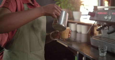 Barista pouring latte with steel pitcher at espresso machine preparing specialty coffee